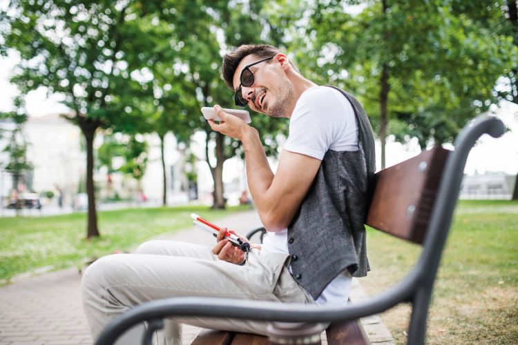 Blinder Mann sitzt im Park auf einer Bank und nutzt sein Smartphone für barrierefreien Zugriff auf Web oder Dokumente